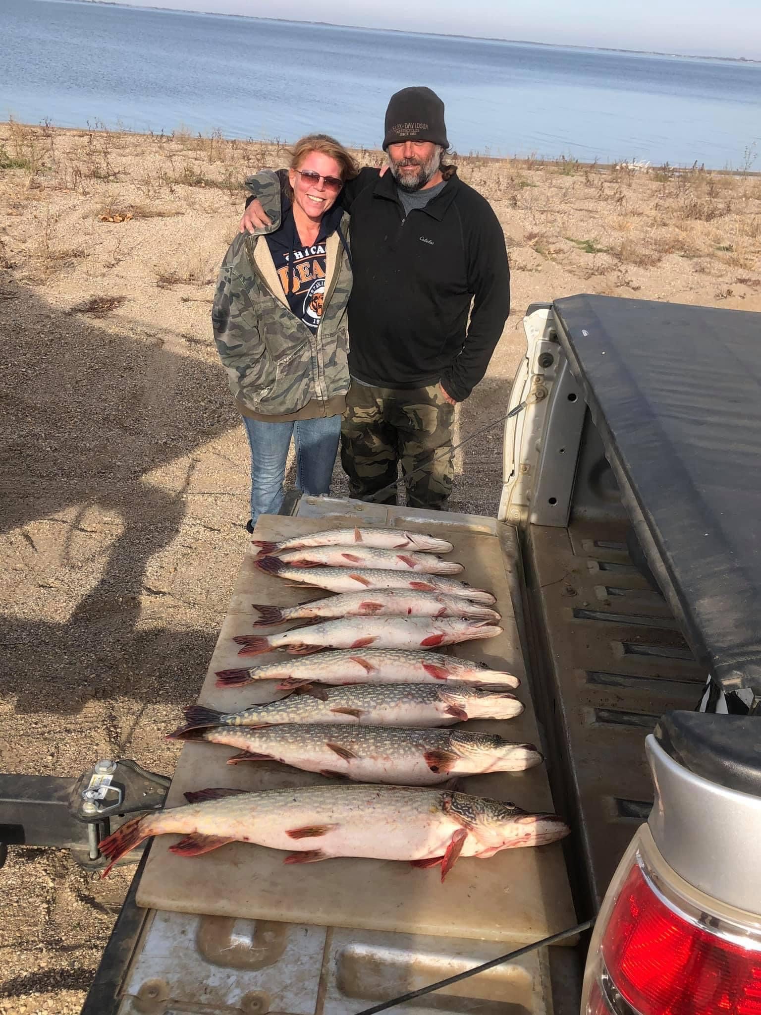 Couple standing near truck bed with a row of caught fish; beach and water in background.