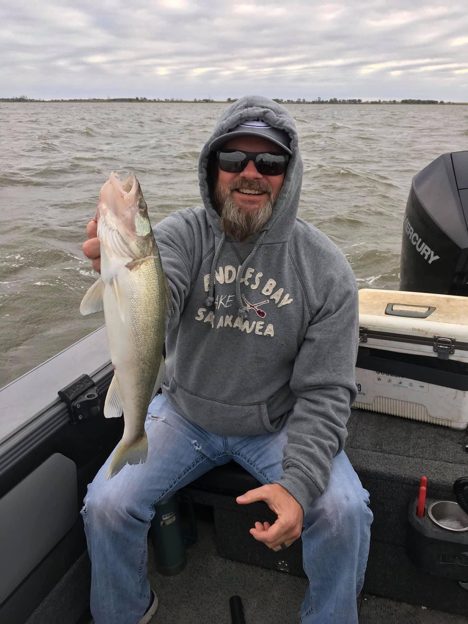 Man in a hooded sweatshirt on a boat holding a fish. Cloudy sky, gray boat, water in the background.