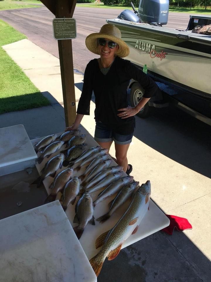Woman with a sun hat poses next to a table laden with a large catch of fish, near a boat.