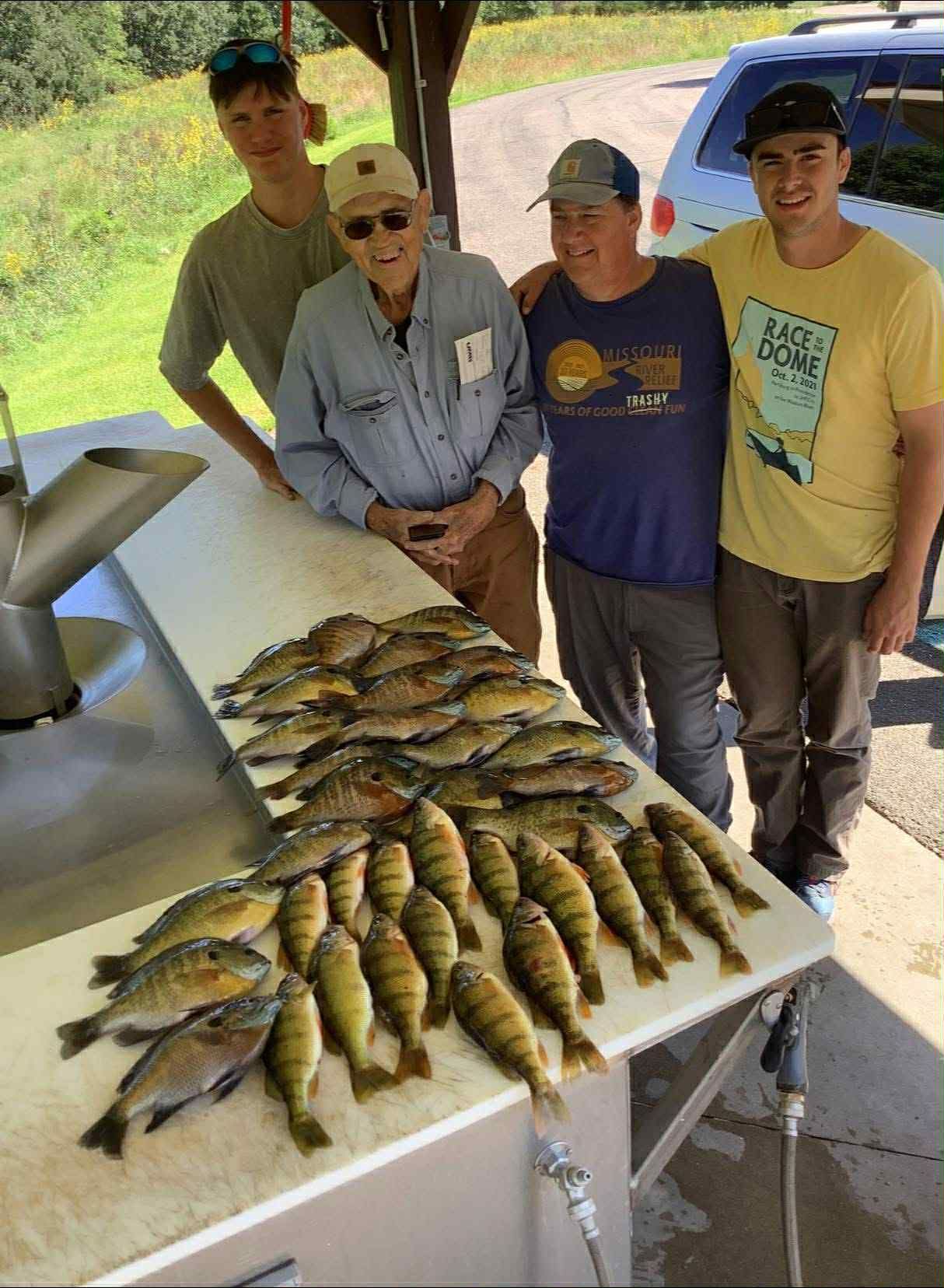 Four men pose with a large catch of fish on a white table under a shelter.