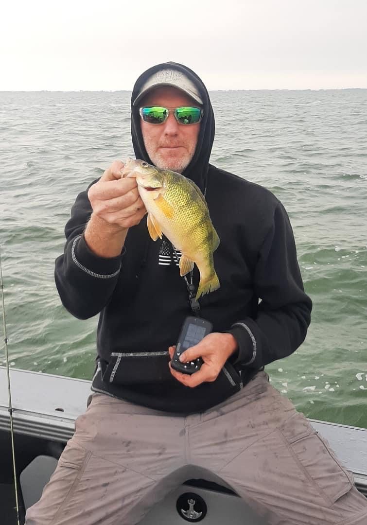 Man in hoodie on boat holding a fish, cloudy water background.