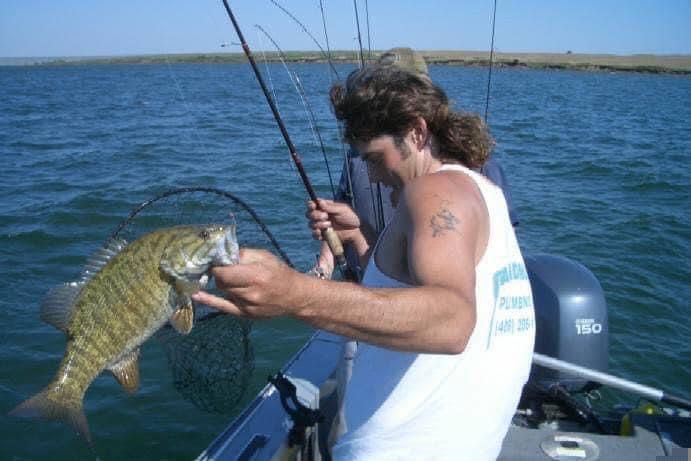 Man on a boat holding a fish, near a net. The lake has a blue color, and the man wears a white tank top.