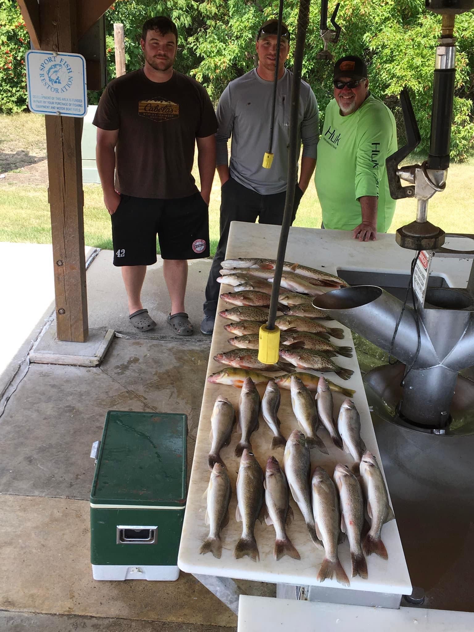 Three men stand behind a table displaying many fish. One man wears sunglasses, another holds a wire. A cooler sits nearby.