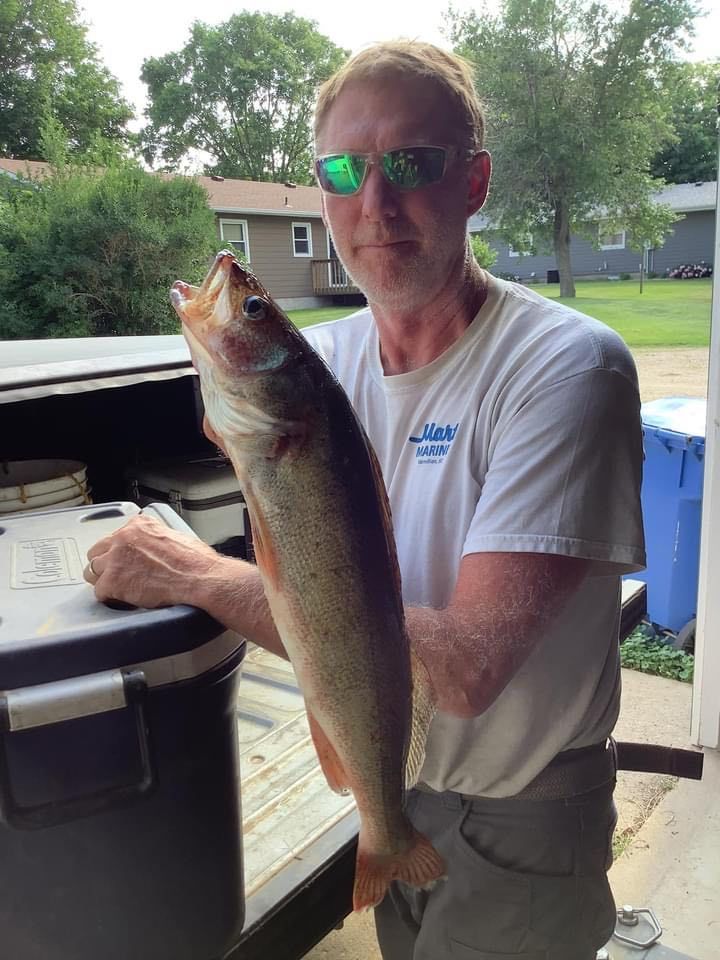 Man holding large fish, standing next to a vehicle. Outdoors, daytime.