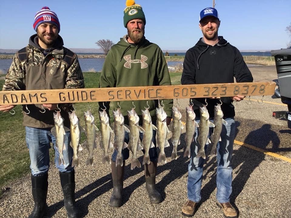 Three men holding up a string of caught fish, posing next to a sign for Adams guide service.