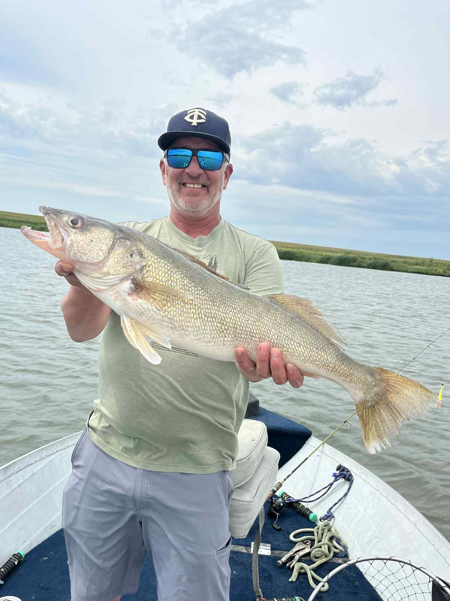 Man on a boat holding a large fish, standing in front of water and green vegetation.