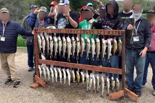 Group displaying many caught fish, held on a wooden rack. People obscured, outdoors.