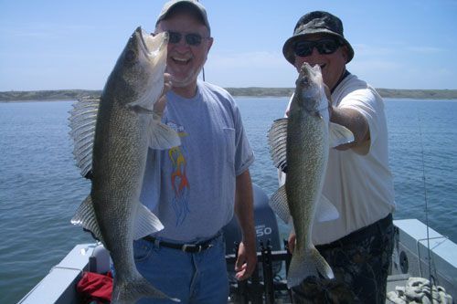 Two men on a boat, holding up large fish, smiling. Blue water and sky in the background.