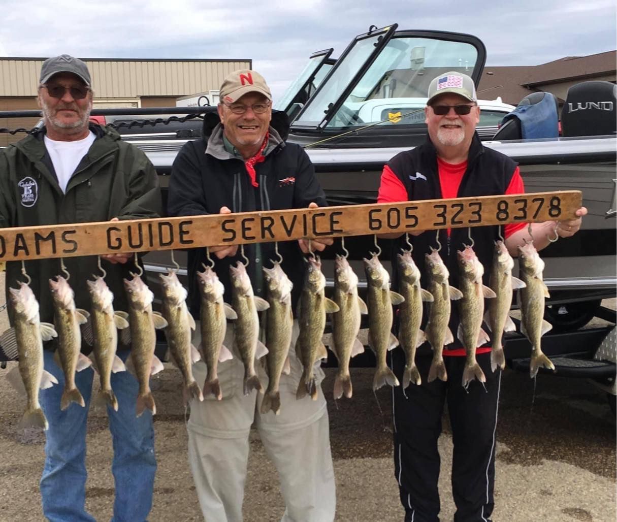 Three men holding a wooden board displaying a string of caught fish near a boat. Board reads “ADAMS GUIDE SERVICE 605 323 8378”.