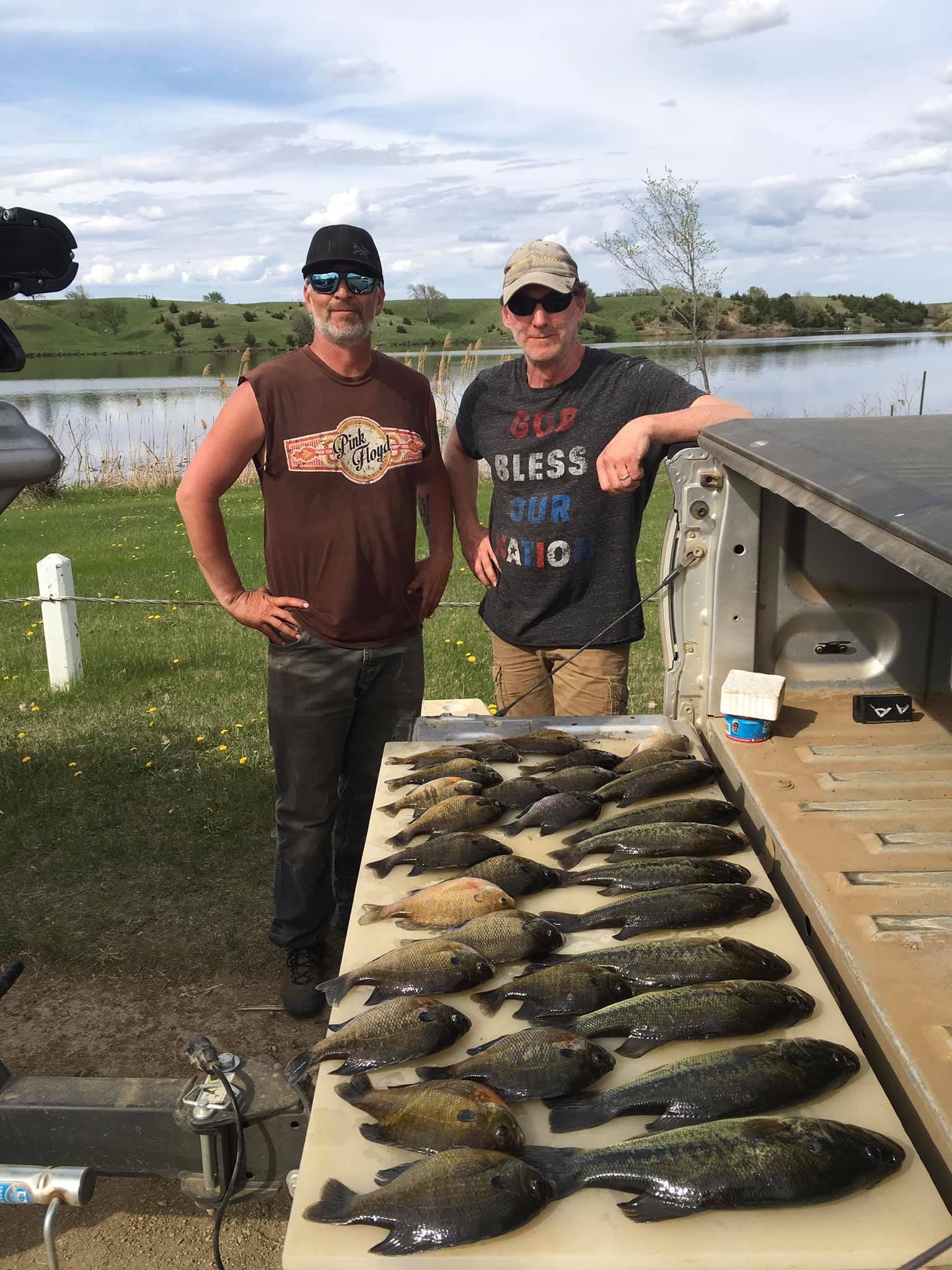 Two men stand near a truck bed displaying many fish. A lake and trees are in the background.
