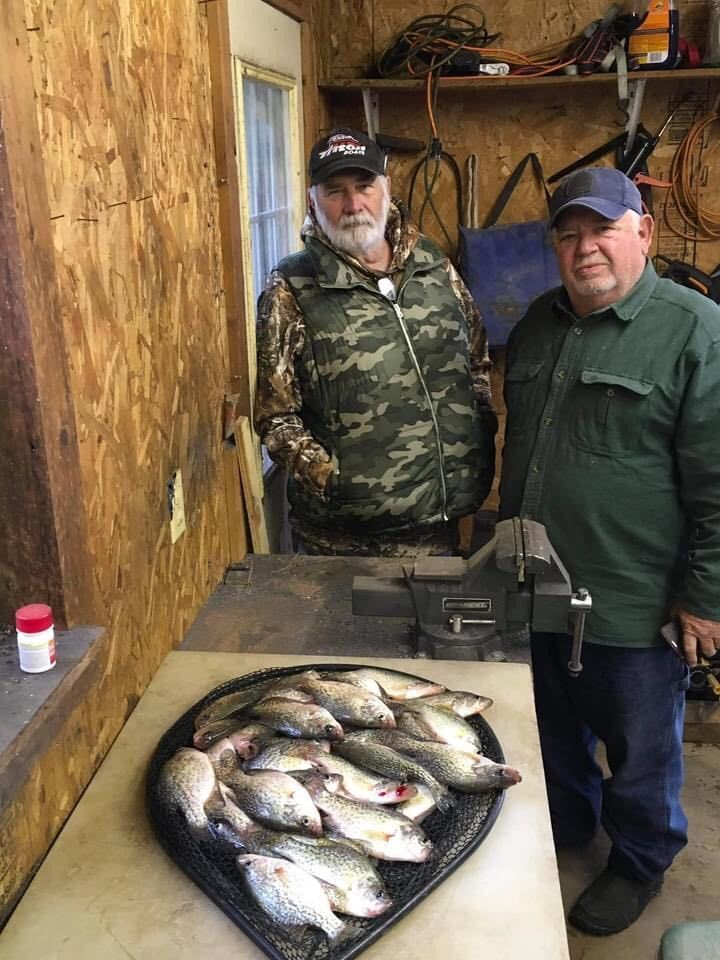 Two men stand behind a table with a pile of fish inside. One wears camo. Wooden garage setting.