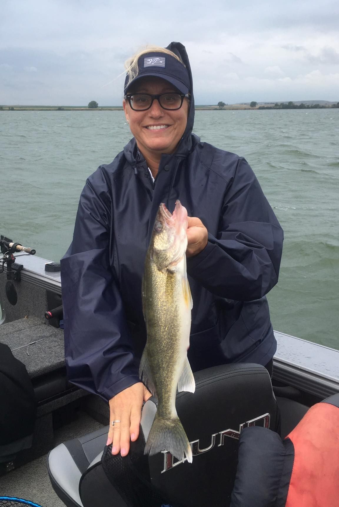 Woman on a boat holding a walleye fish. Wearing glasses, a hat, and a blue jacket on a cloudy day.