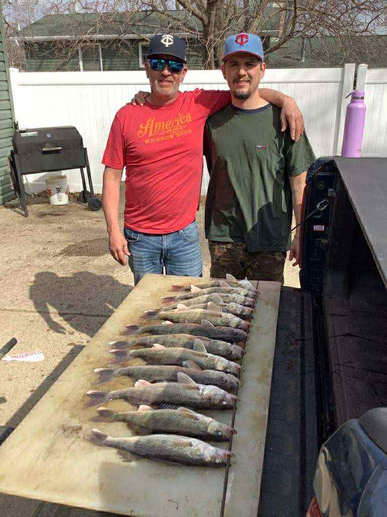 Two men pose with a row of caught fish on a cutting board outdoors.