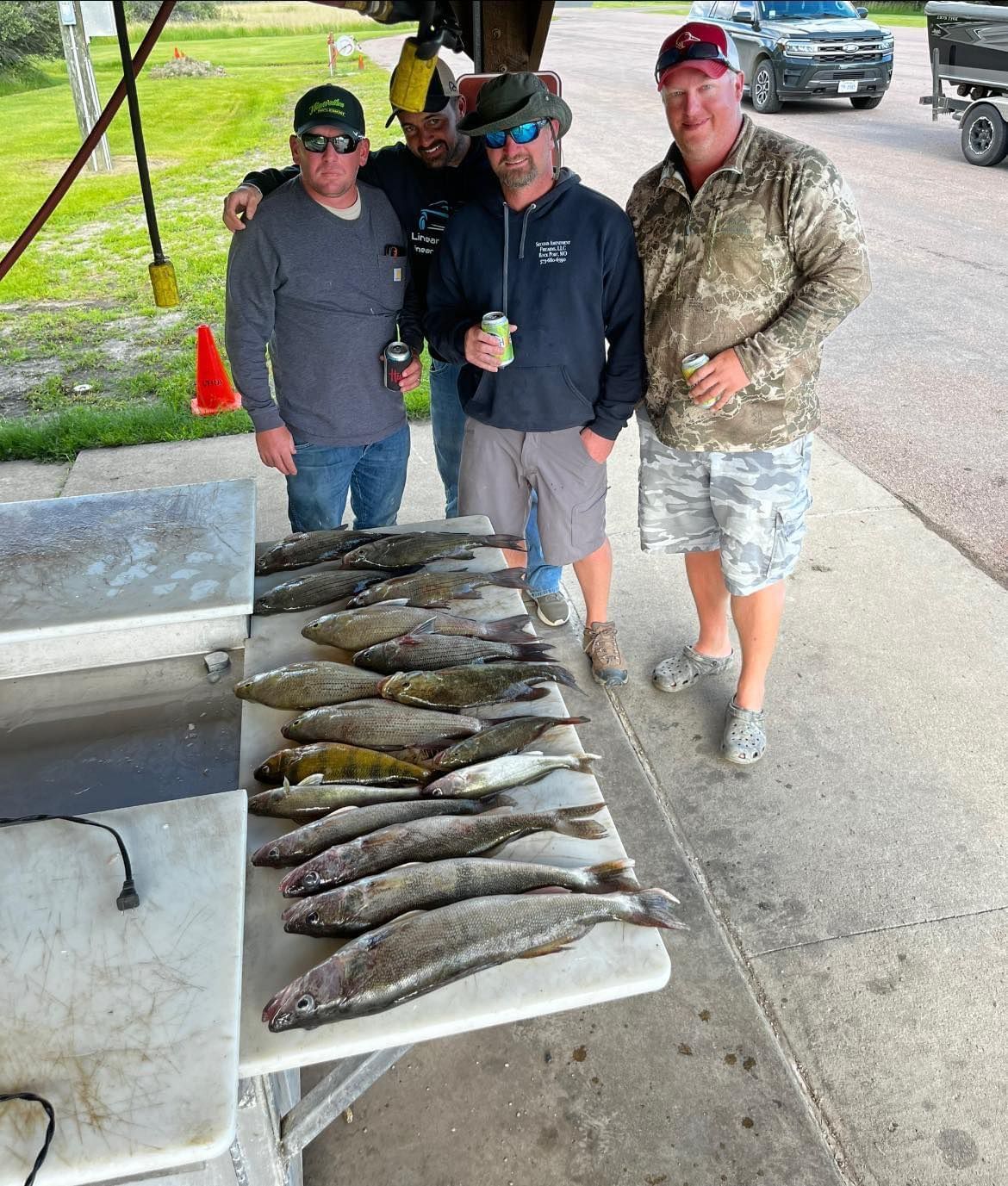 Four men pose with a table of fish; some wear hats and sunglasses. Outdoor setting.