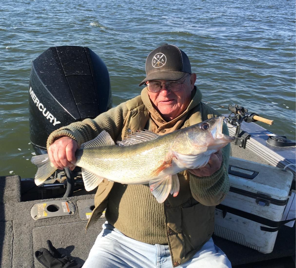 Man holding up a walleye fish on a boat. Black motor visible, calm water, overcast day.