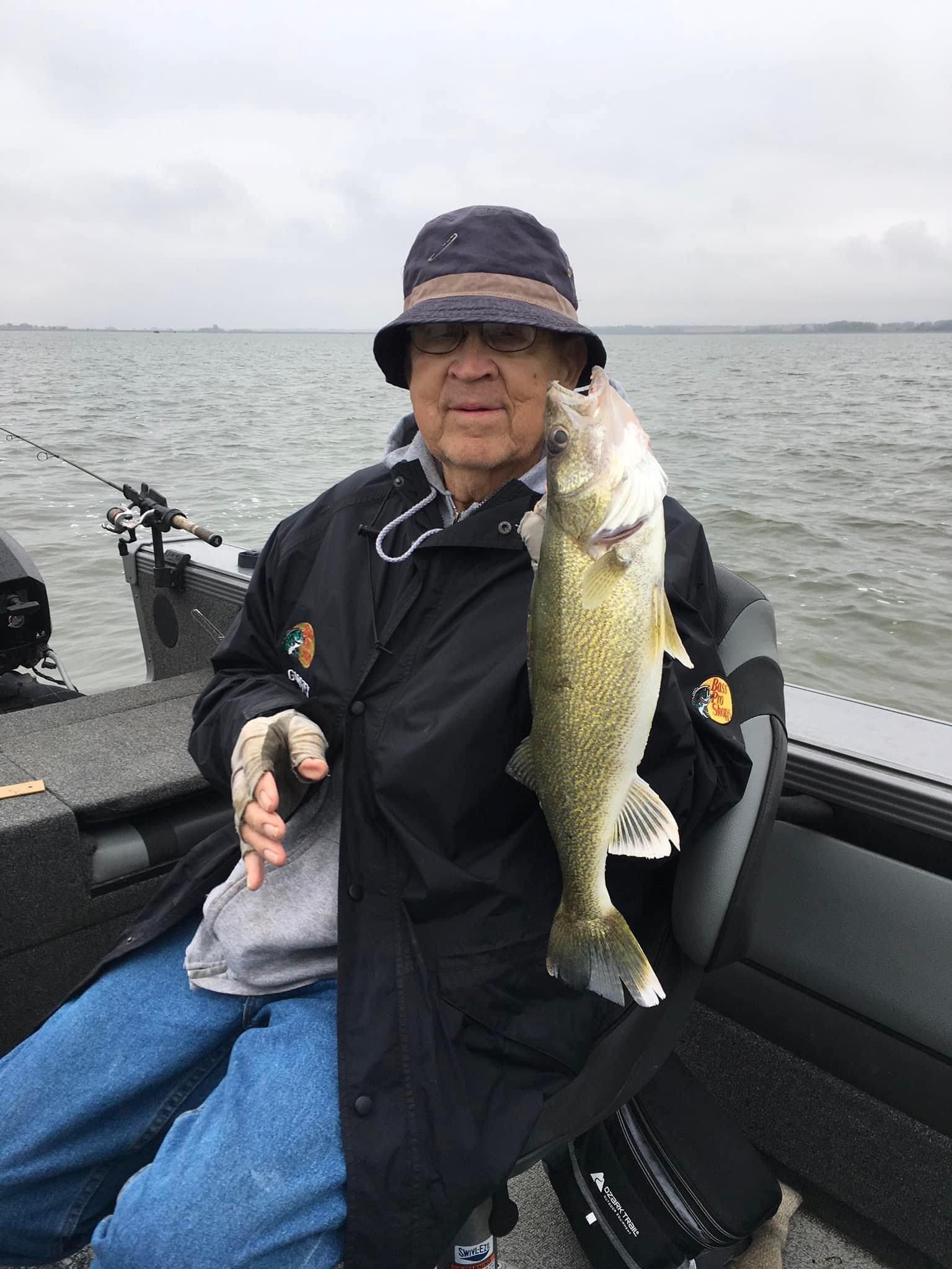 Man in a fishing boat holding up a walleye. Overcast sky, lake in the background.