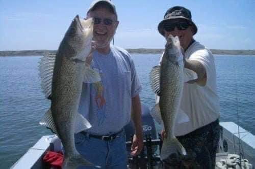 Two people on a boat holding up large fish they caught, smiling, with a lake and blue sky in the background.