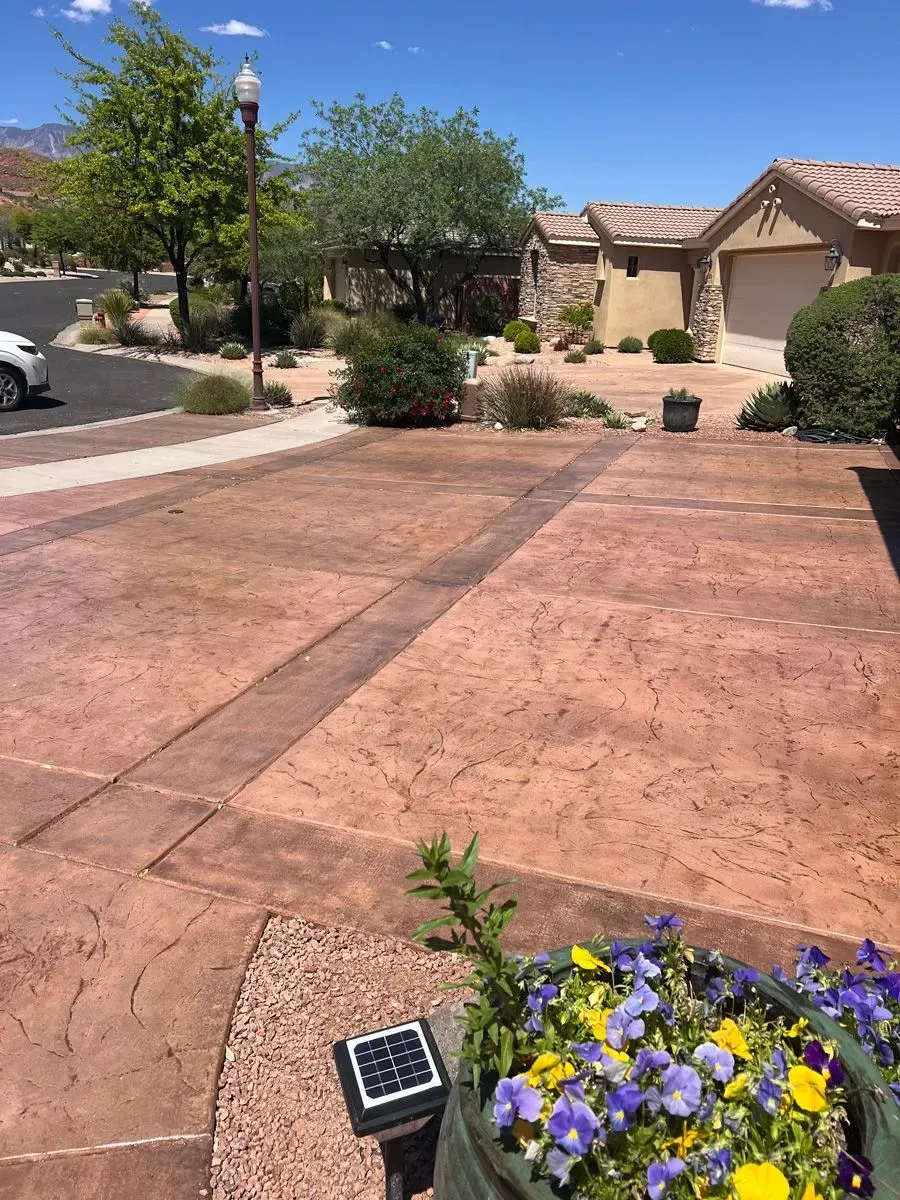 Driveway leading to a house, featuring a patterned, red-toned concrete surface. Blue and yellow flowers in the foreground.