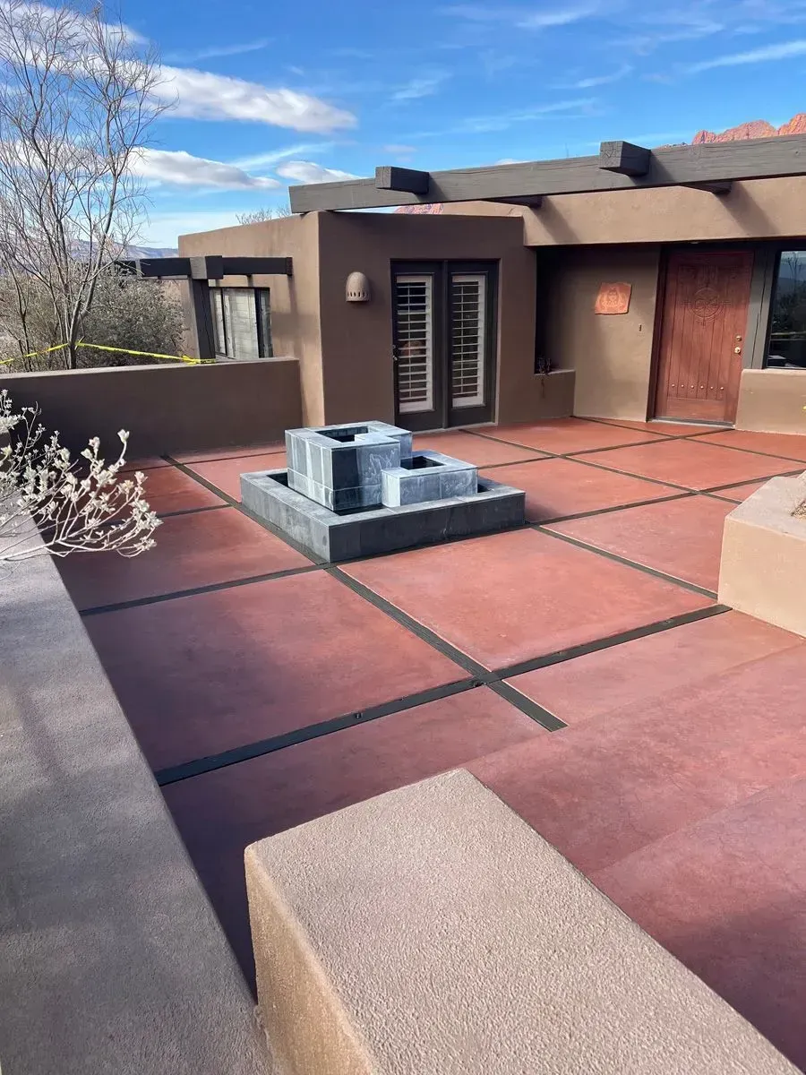 Red patio with fountain, dark lines, and brown building. Blue sky.