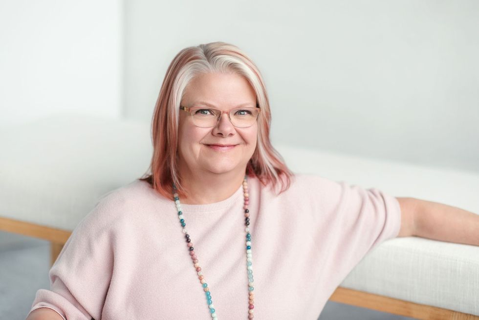 A close up head shot of a blond woman in a pink sweater with colorful jewelry and glasses