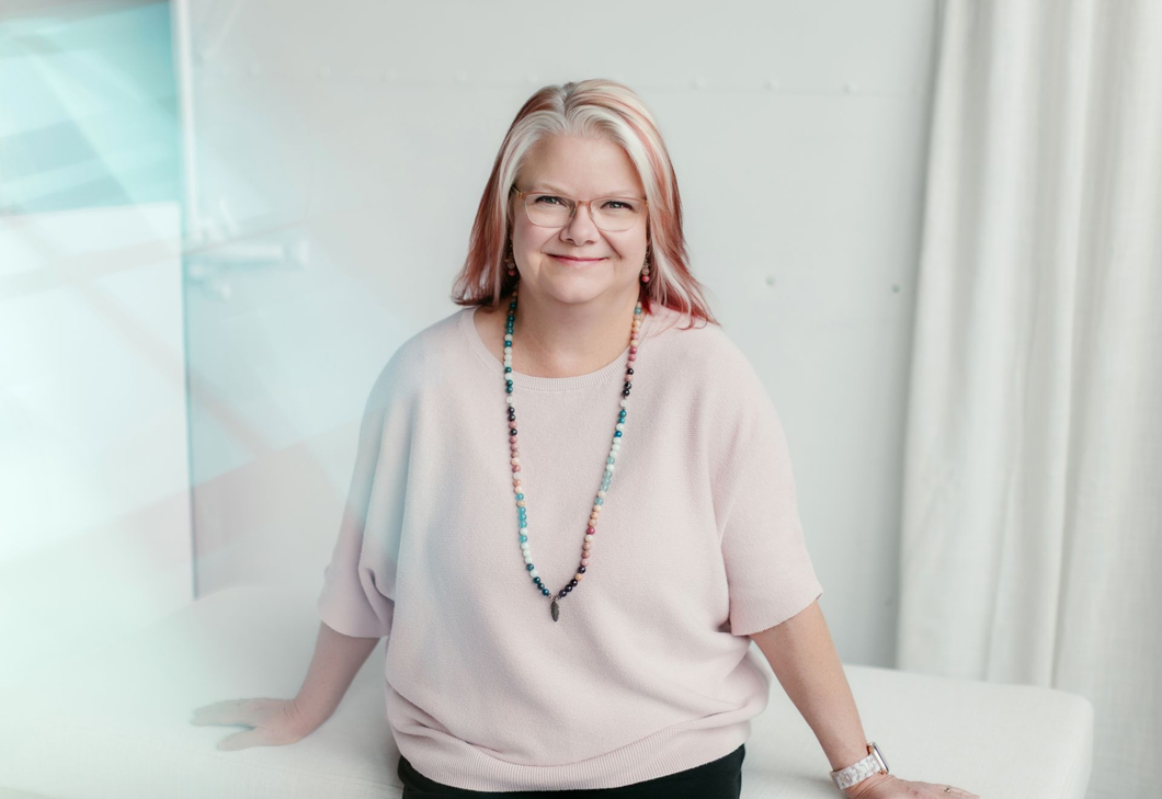 Woman in a light pink top smiling indoors beside a white curtain