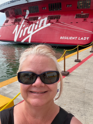 A woman in sunglasses smiling in front of the  stern of Virgin Voyages ship, Resilient Lady