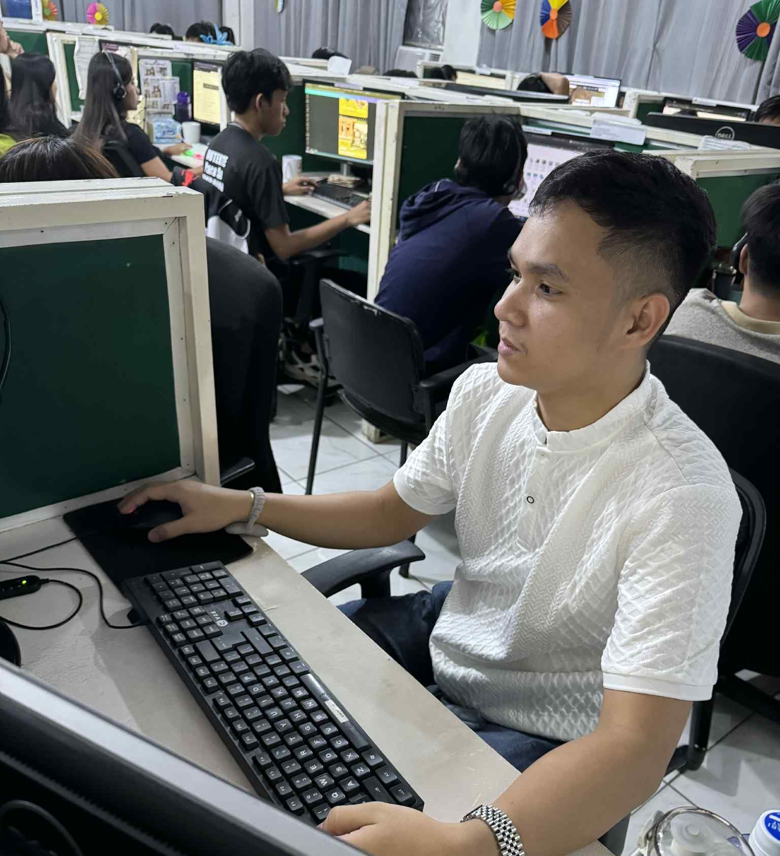 Man working on a computer in a busy office setting. He is wearing a white shirt and using a mouse and keyboard.