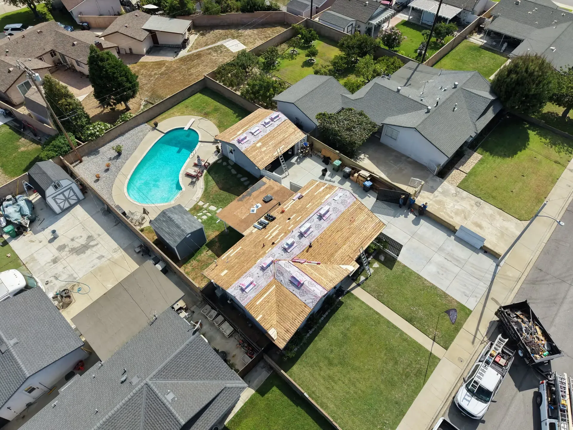 Aerial view of a suburban home under roof repair beside a backyard pool and neighboring houses.