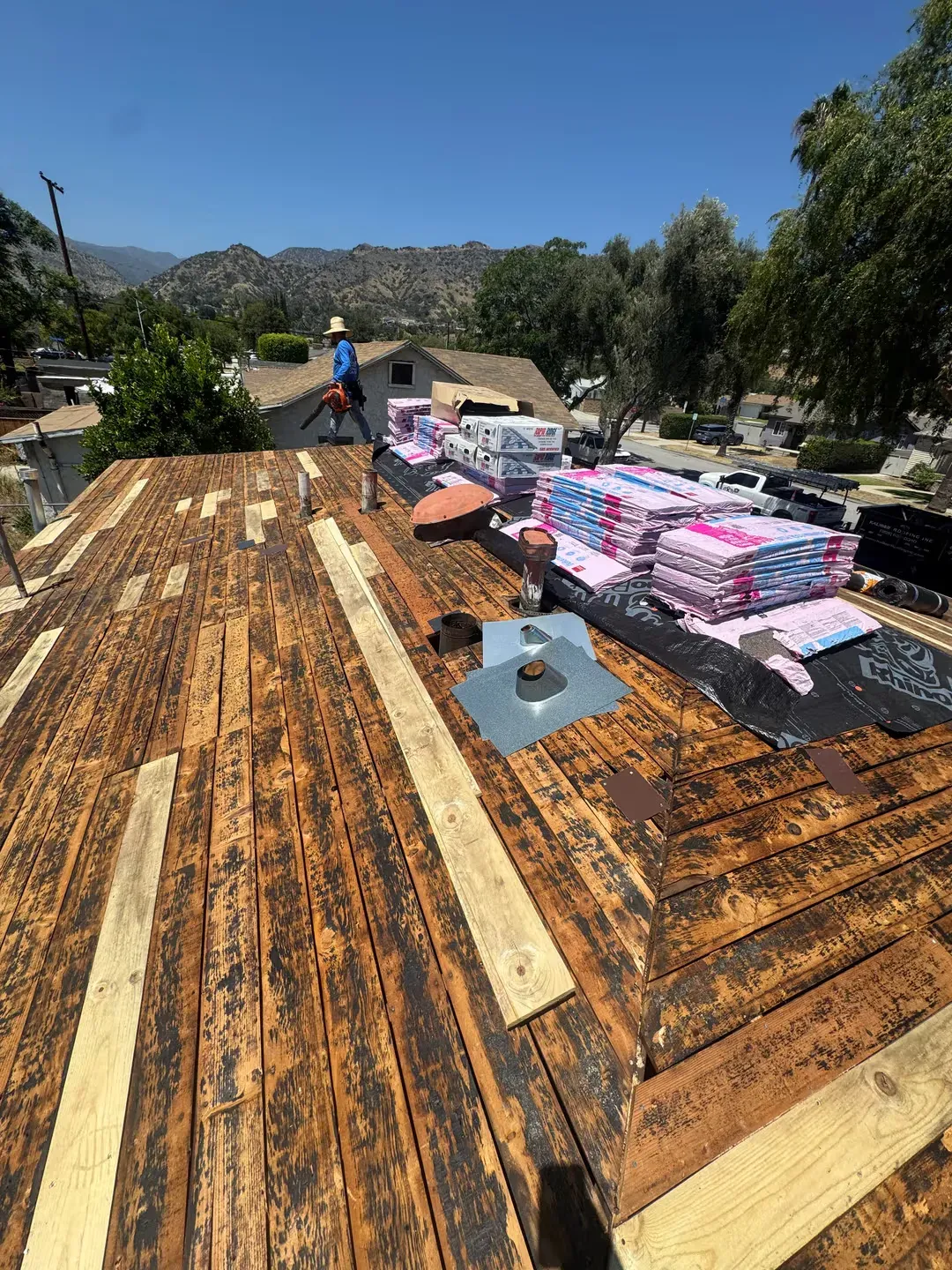 Workers install roofing on a house, with stacks of shingles on a sunlit roof under a clear sky.