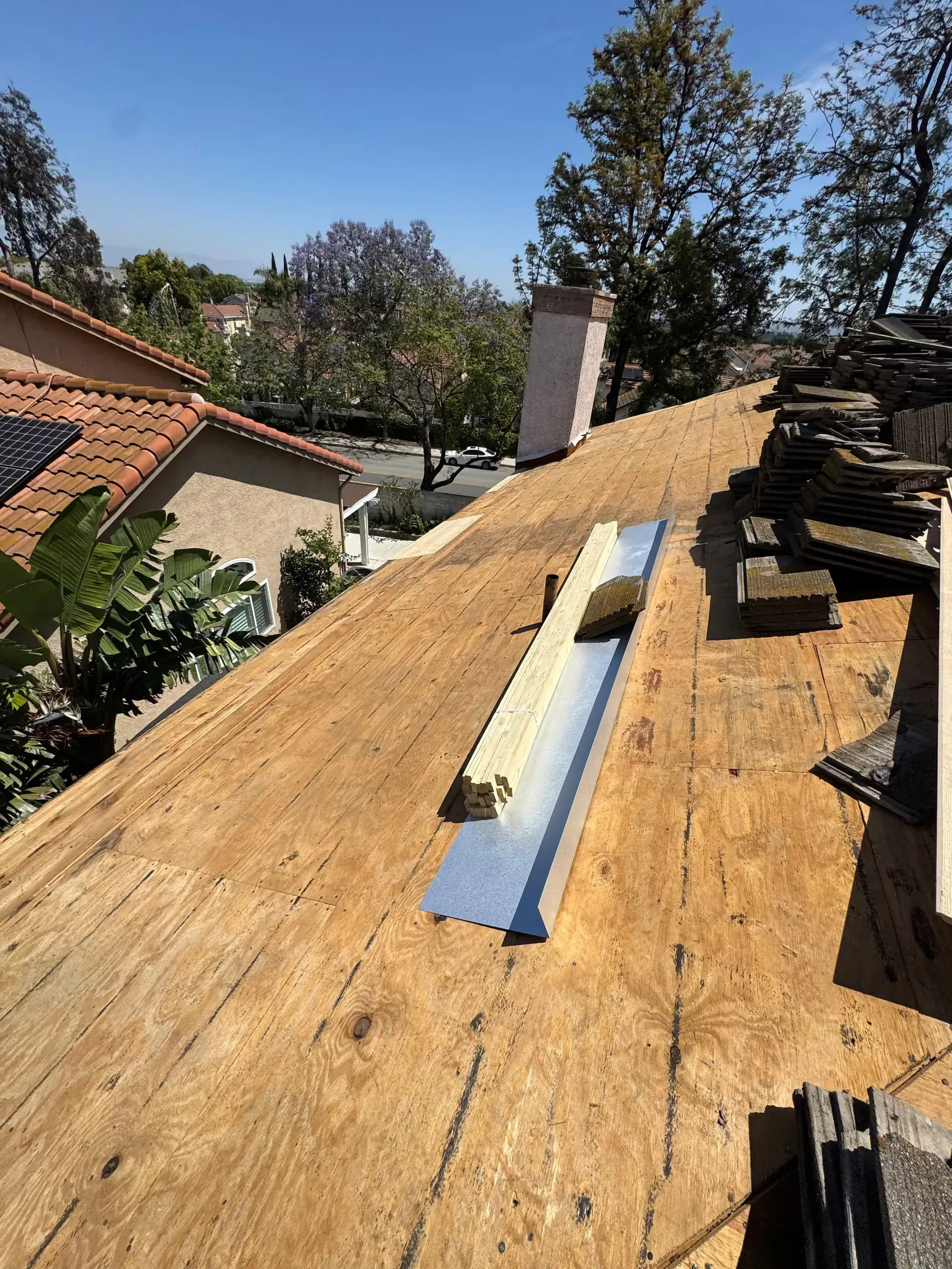 Roof under construction with plywood sheathing, chimney, and stacked shingles beside neighboring houses