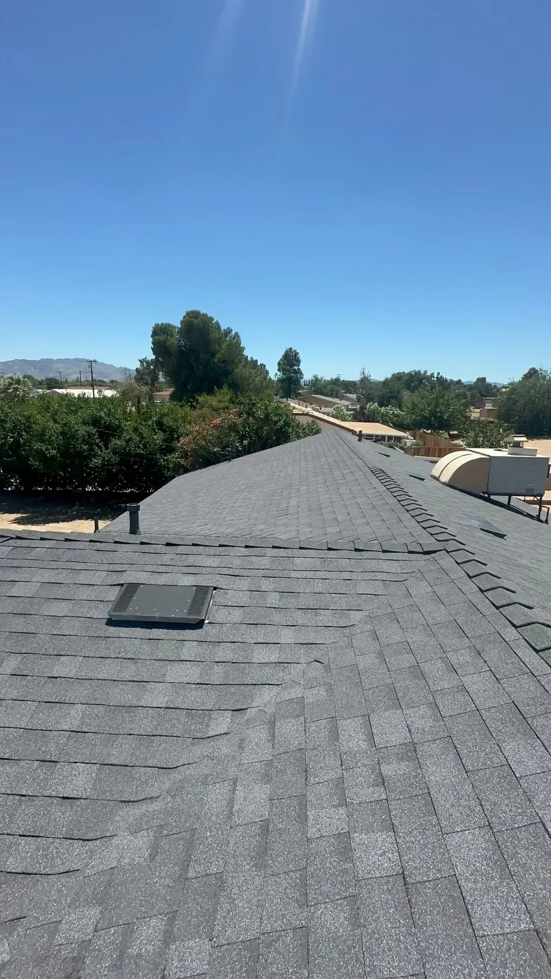 Rooftop with gray shingles under a clear blue sky, bordered by trees and distant hills