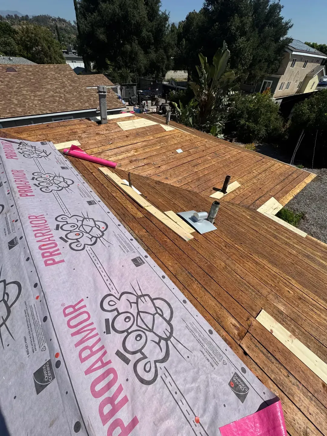Roof under construction with exposed plywood, pink underlayment, and scattered tools in a sunny neighborhood.