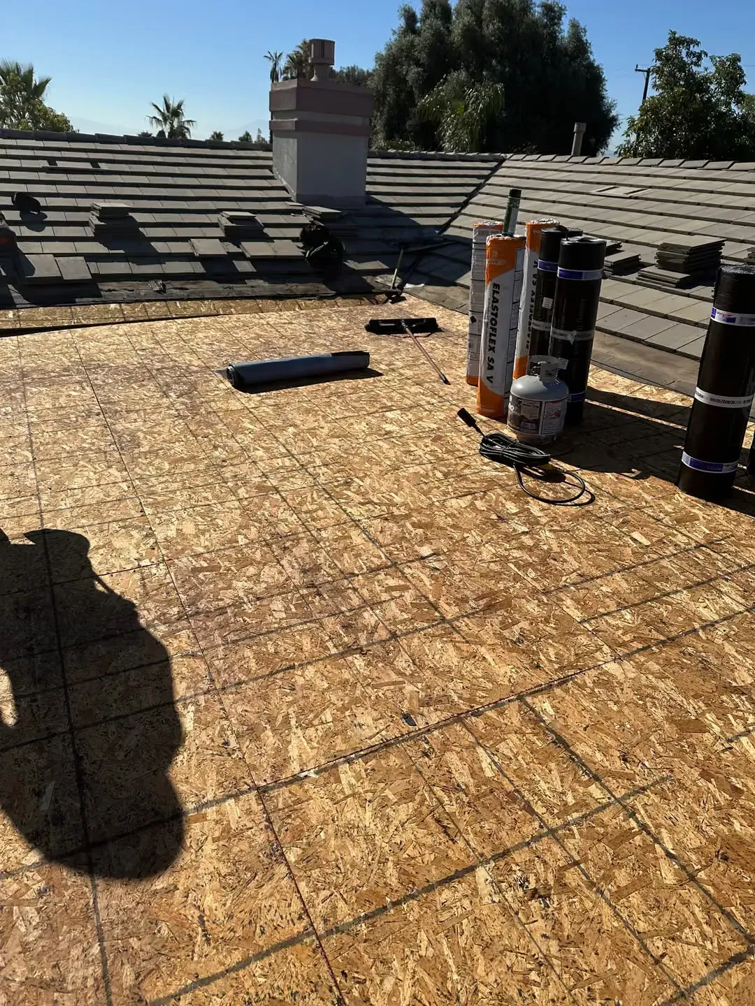 Flat roof under construction with exposed plywood decking, roof vents, and rooftop equipment in bright sunlight