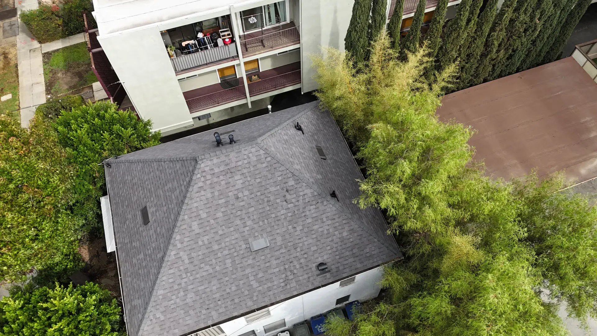 Aerial view of a gray-roofed house beside a taller building and dense green trees.