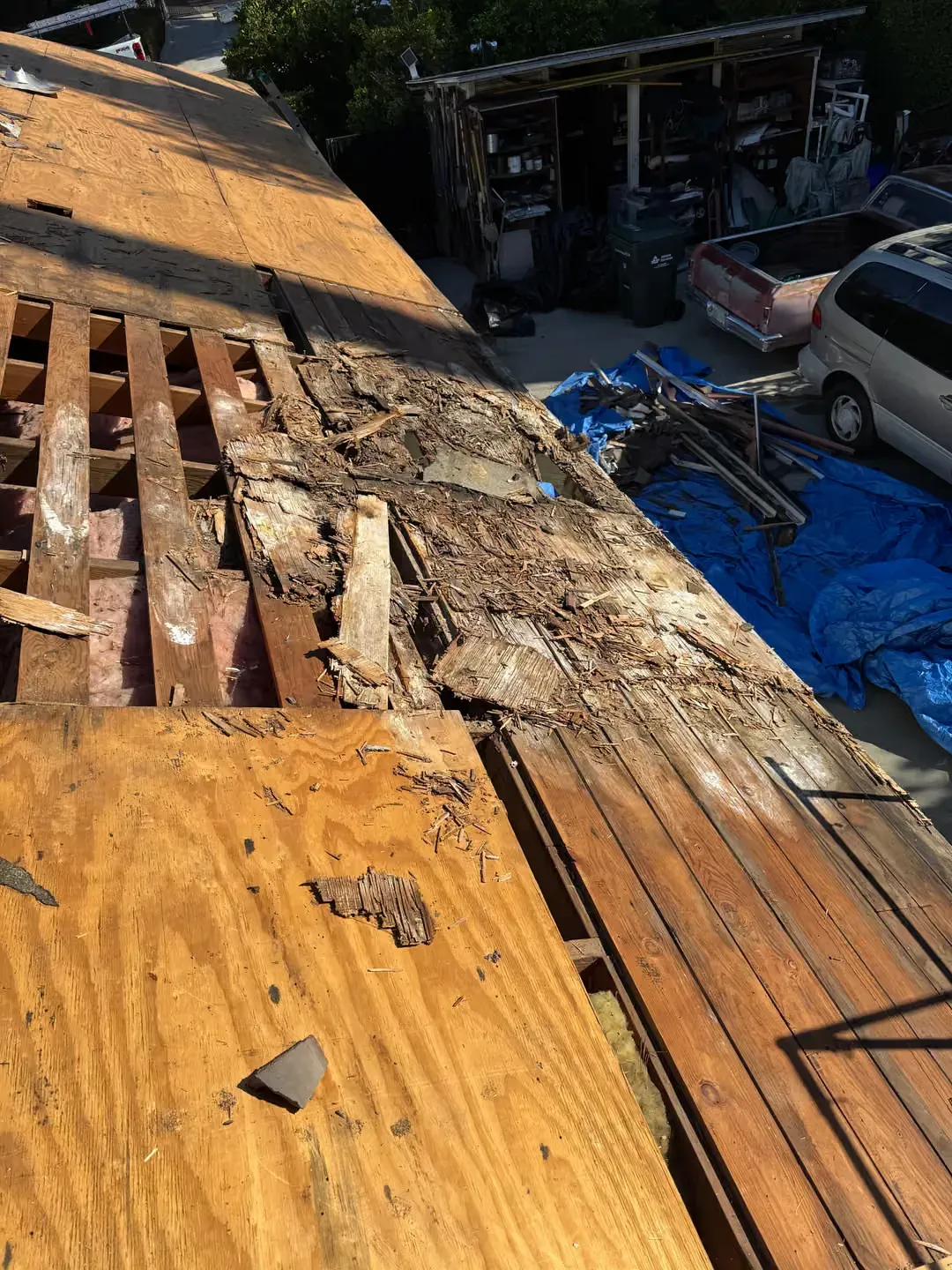 Damaged roof with exposed beams and torn shingles beside a driveway and parked car