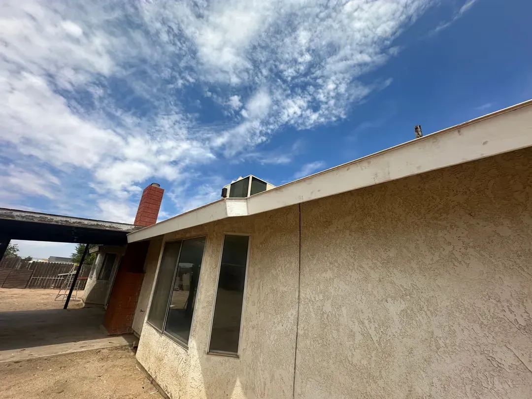 Single-story stucco house with tan walls, a red brick chimney, and blue sky with clouds overhead
