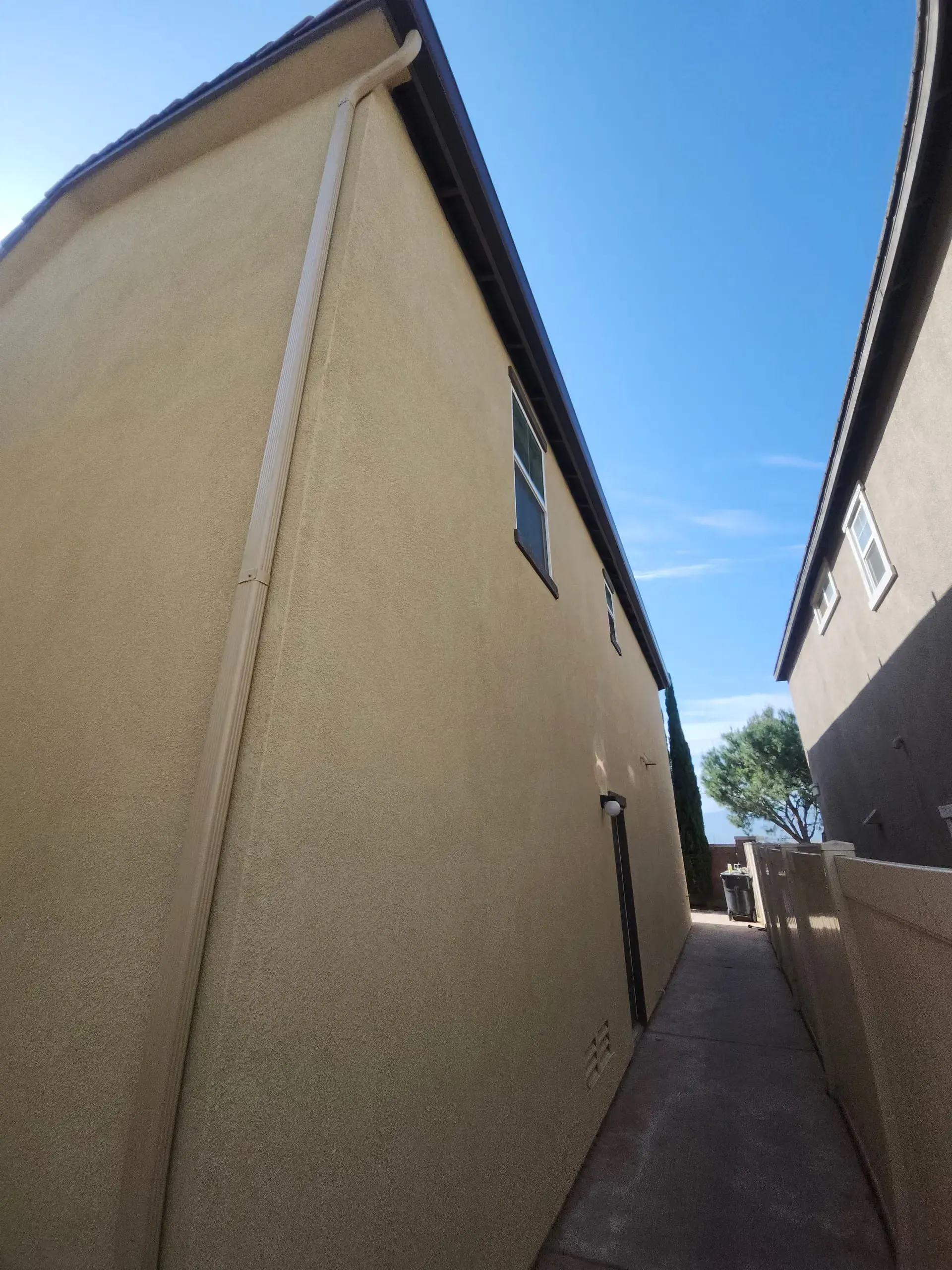 Narrow alley between beige stucco houses under a clear blue sky.