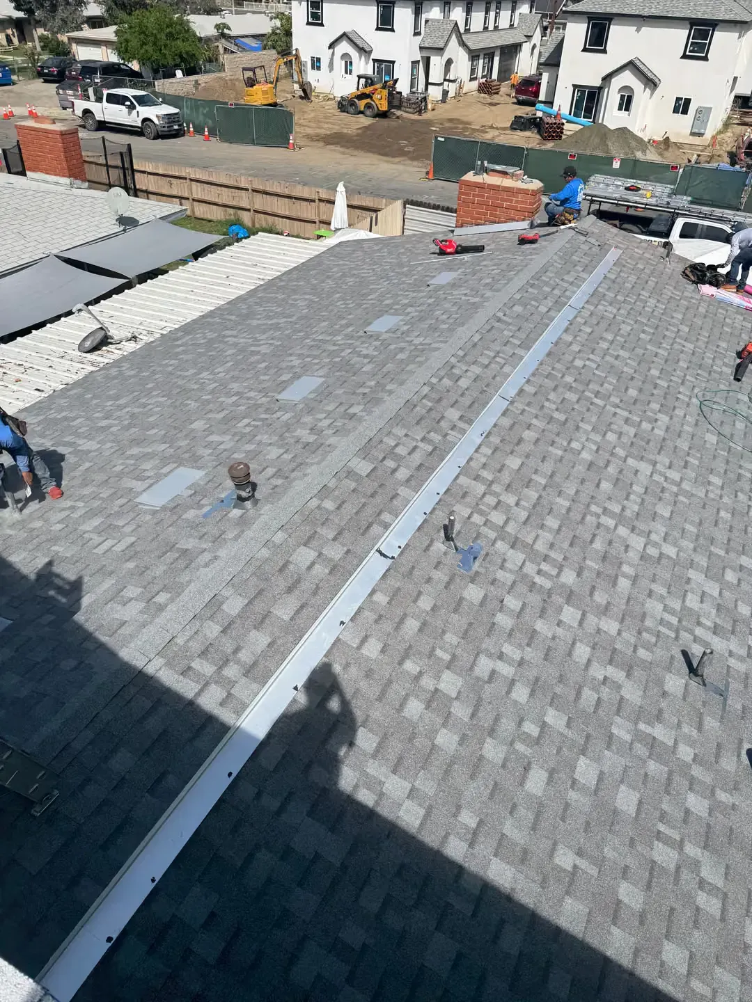 Rooftop with gray shingles and white trim under construction, seen above houses and parked cars.