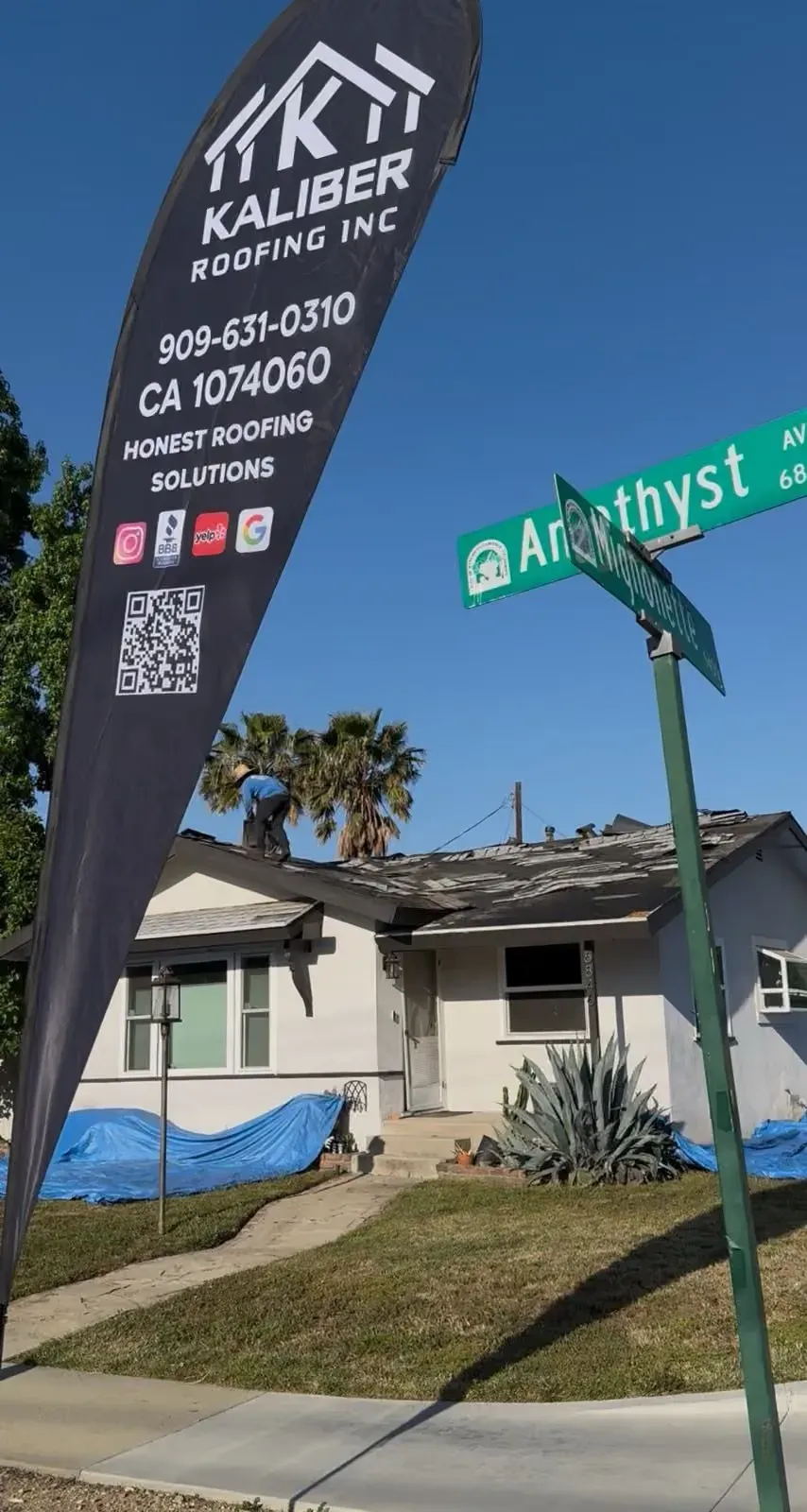 Kaliber Roofing sign on a house corner by street sign and blue tarp under a clear sky