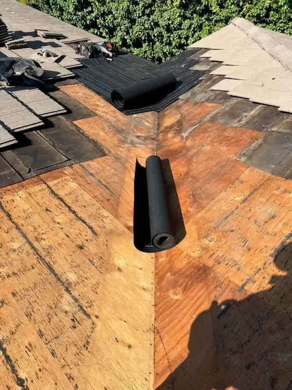 Roof valley under repair with orange underlayment and black roofing rolls near exposed shingles
