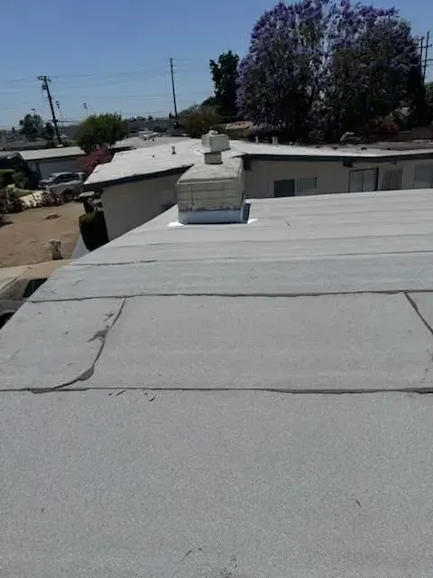 Flat gray rooftop with a small chimney, cracks, and houses and trees in the background