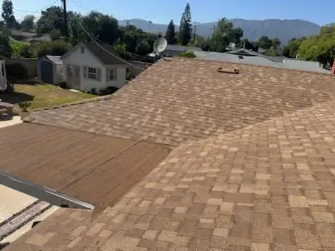 Roof under construction with brown shingles and a partial section of exposed plywood, suburban homes in background
