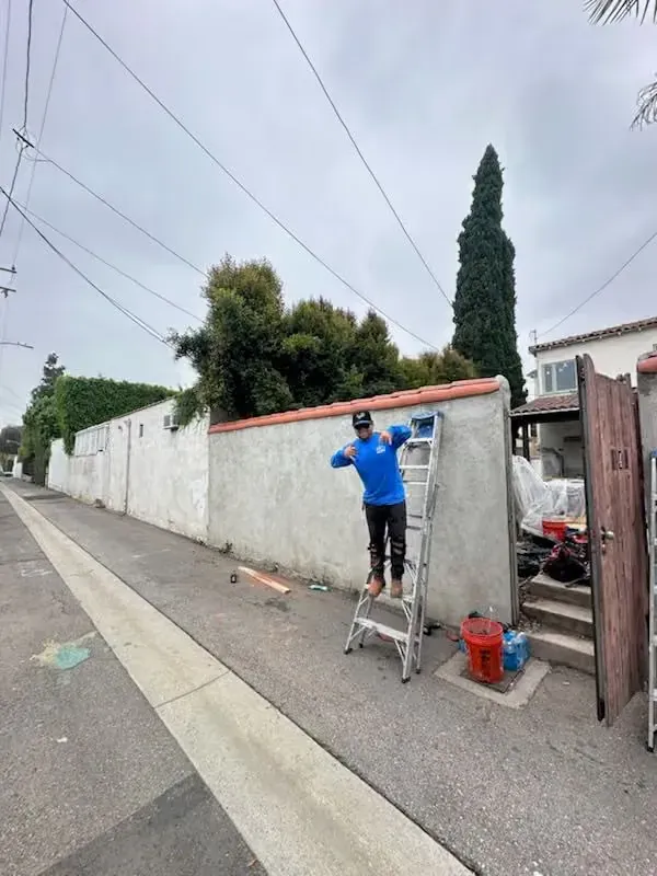 Worker in blue jacket stands on a step ladder painting a concrete wall beside a sidewalk.