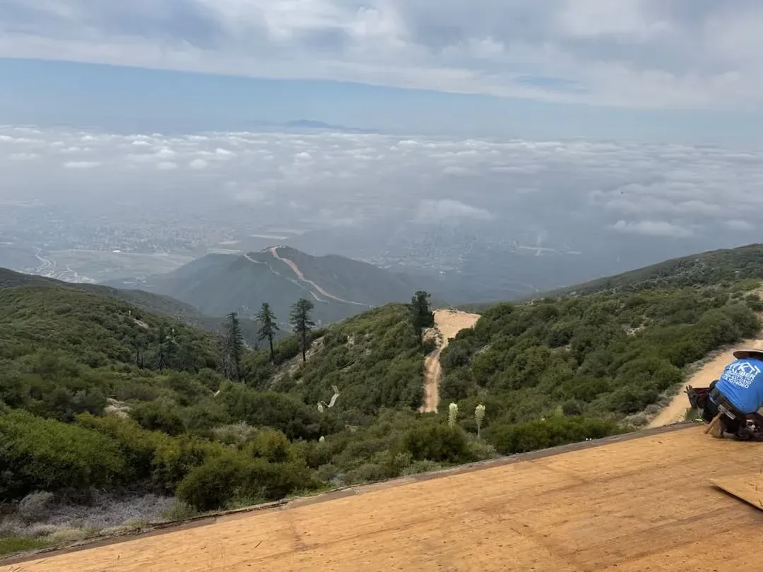 Mountain trail overlooking a hazy valley, with green hills, a dirt path, and a blue sign on the right.