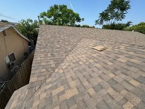 Roof covered in tan shingles, with a small roof hatch and trees visible in the background.