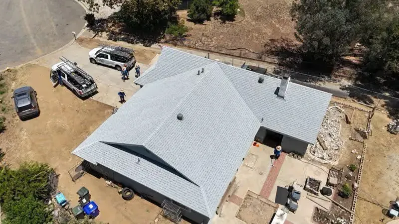 Aerial view of a gray-roofed house with parked vehicles and construction materials in a dusty yard.