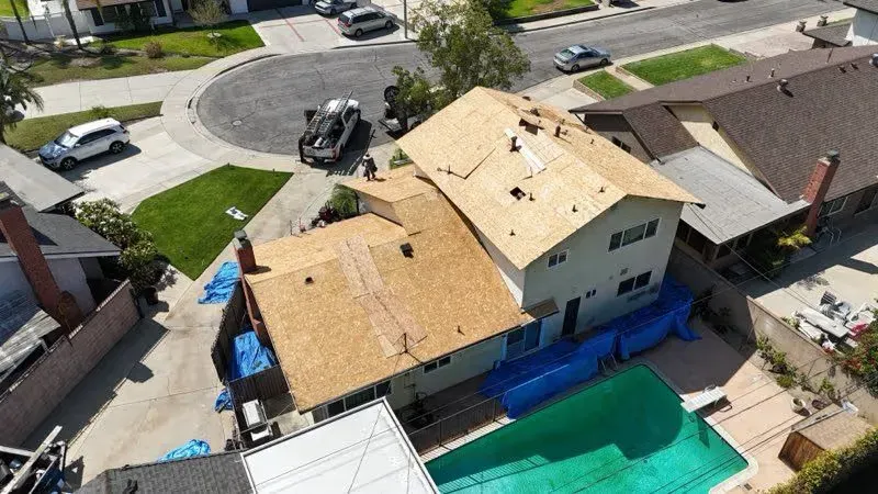 Aerial view of a house with a damaged roof, surrounded by blue tarps and neighboring homes.