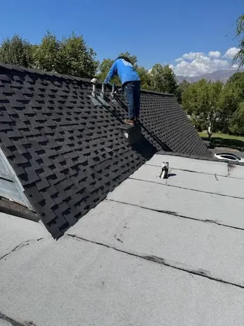 Worker installing shingles on a steep black roof beside a flat white roof under a blue sky