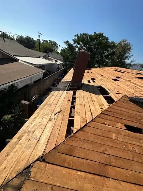 Roof under repair with exposed wooden decking and a brick chimney, seen from above on a sunny day