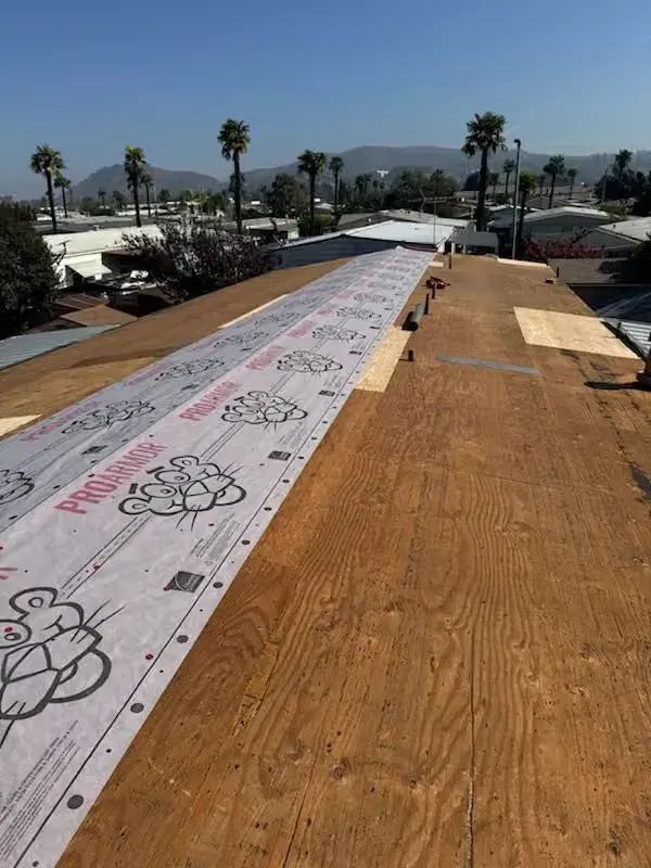 Rooftop under construction with white safety wrap and brown plywood decking, palm trees and mountains in the background